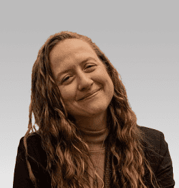 A person with long, wavy hair wearing a dark jacket and turtleneck smiles at the camera against a plain gray background.