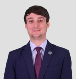 A man in a suit with a maroon tie and a pin on his lapel stands against a plain background.