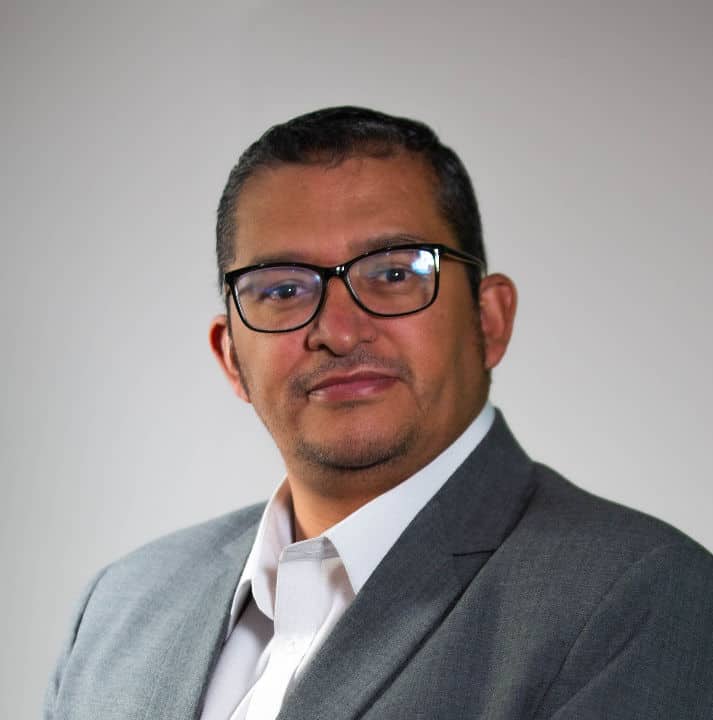 Person wearing glasses and a gray suit with a white shirt, looking at the camera against a neutral background.