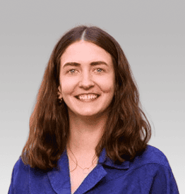 A woman with shoulder-length brown hair, wearing a blue collared shirt, smiles at the camera against a gray background.