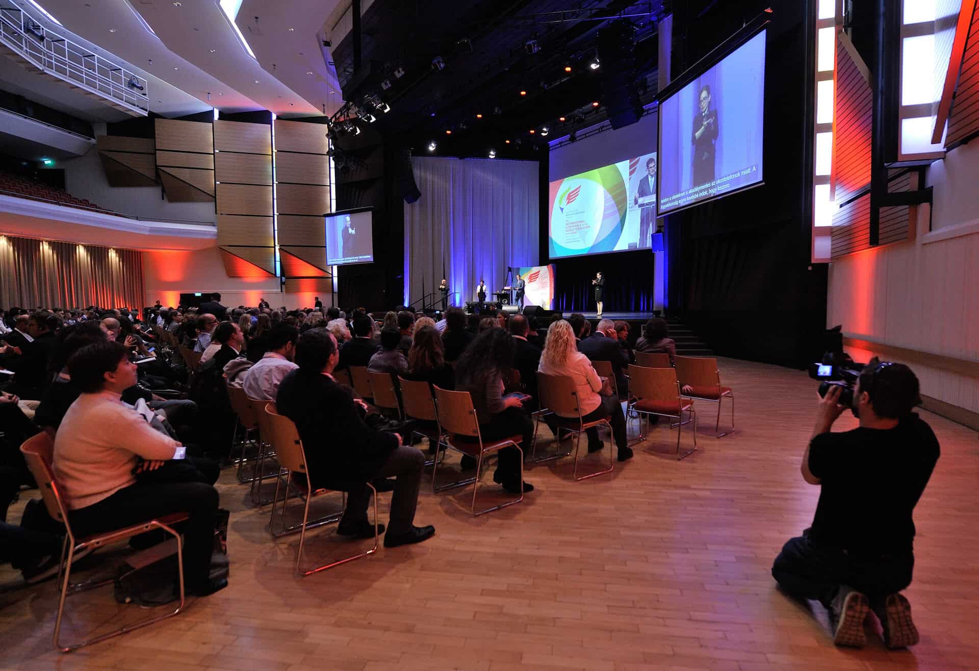 Audience seated in a large auditorium watching a presentation on stage, with a cameraman capturing the event.