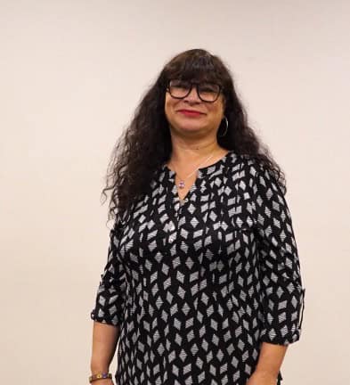 A woman with long curly hair and glasses stands smiling, wearing a black and white patterned dress against a plain background.