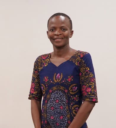 A person stands against a plain background, wearing a blue dress with pink and yellow floral patterns.