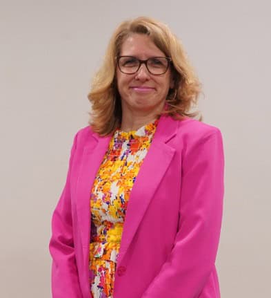 A person with glasses wearing a pink blazer and colorful dress stands against a plain background.