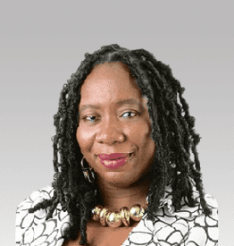 A person with long braided hair and a patterned outfit, wearing large earrings and a chunky necklace, smiles slightly against a gray background.