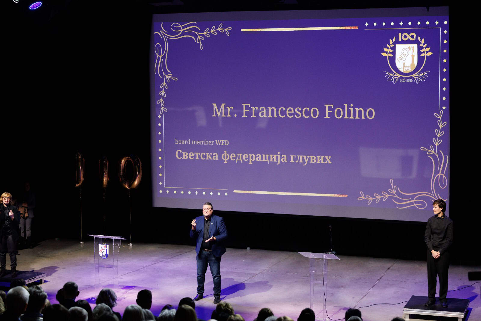A man stands on stage speaking with a presentation slide behind him displaying "Mr Francesco Folino" and Cyrillic text at an indoor event.