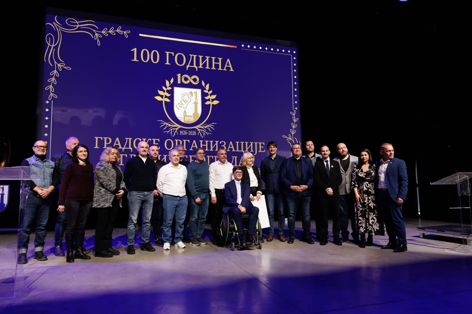 A group of people pose on stage in front of a large screen displaying "100 godina" and an emblem, marking a centenary celebration event.