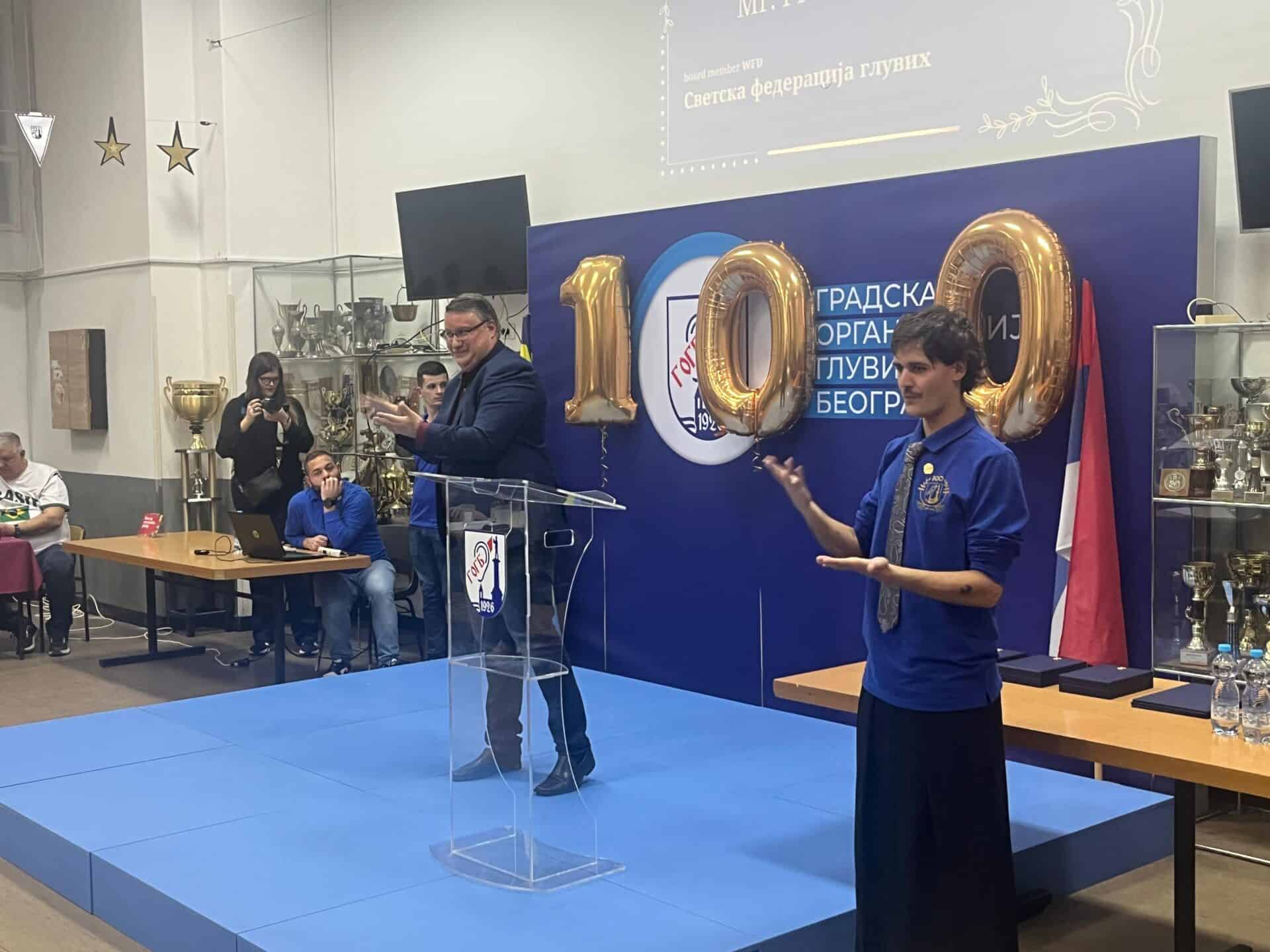 A man speaks at a lectern next to a woman doing sign language, with gold "100" balloons and trophies in the background at an indoor event.