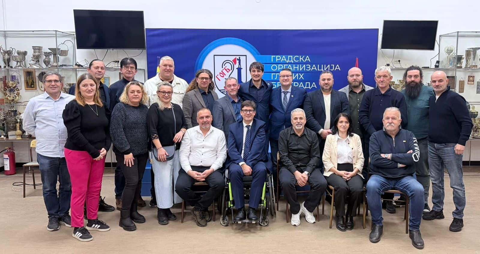 A group of 21 adults pose for a group photo indoors, standing and sitting in front of a blue banner with Serbian text and surrounded by trophy displays.