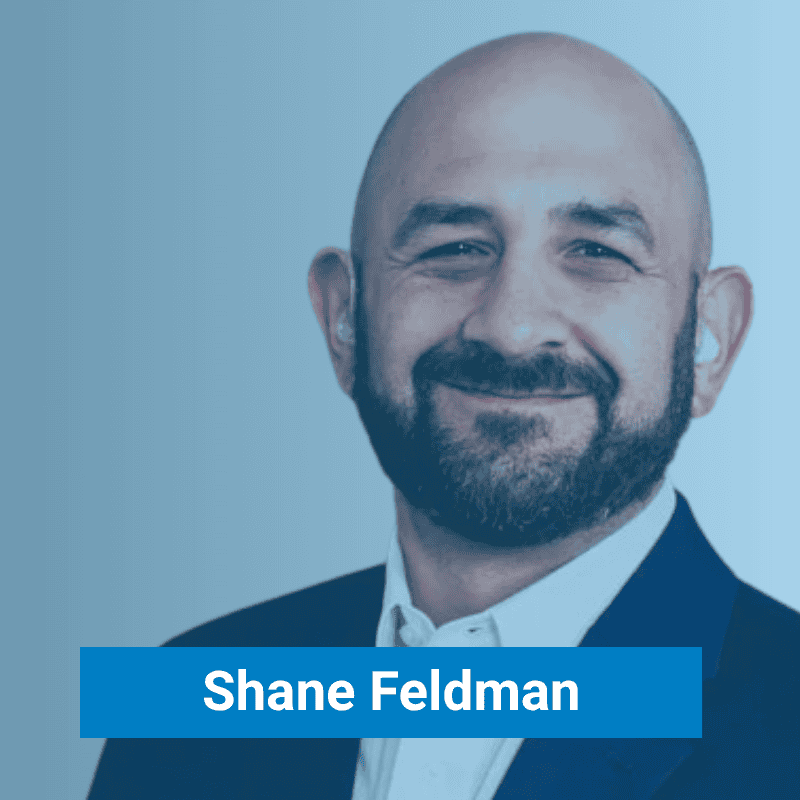 A bald man with a beard wearing a suit and white shirt is smiling at the camera. The name "Shane Feldman" appears in white text on a blue label at the bottom.