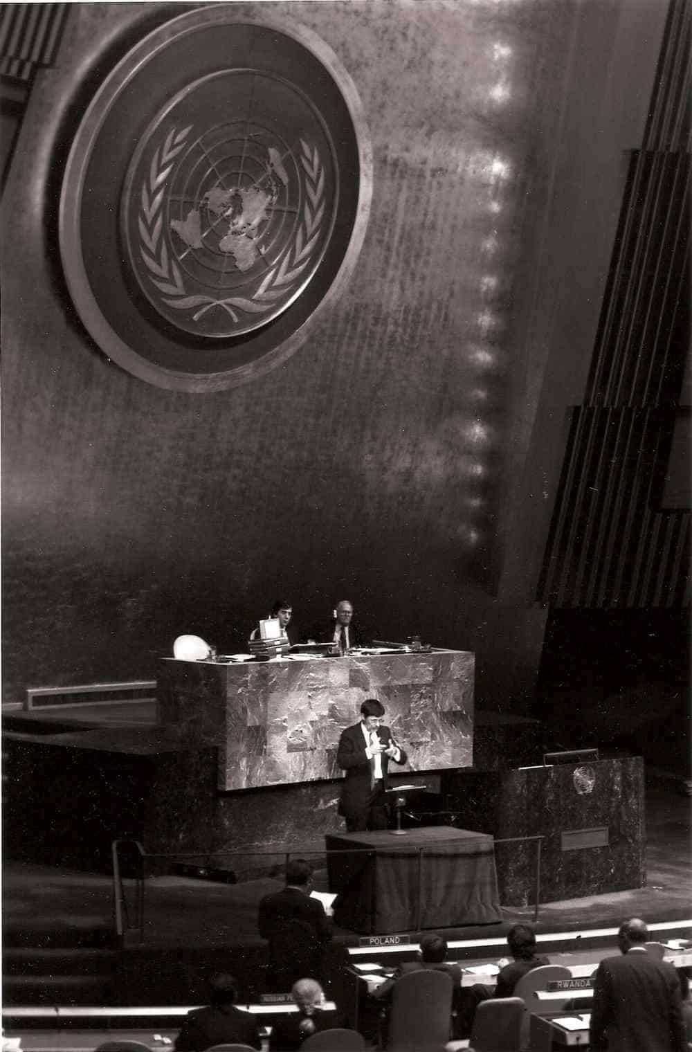 A man stands at a podium speaking in a large hall with the United Nations emblem visible behind him.