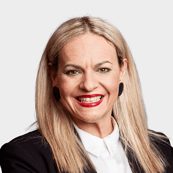 Smiling person with long blonde hair, wearing a black blazer and white shirt, against a plain background.