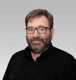 A man with glasses, a beard, and short hair, wearing a black shirt, poses against a gradient gray background.