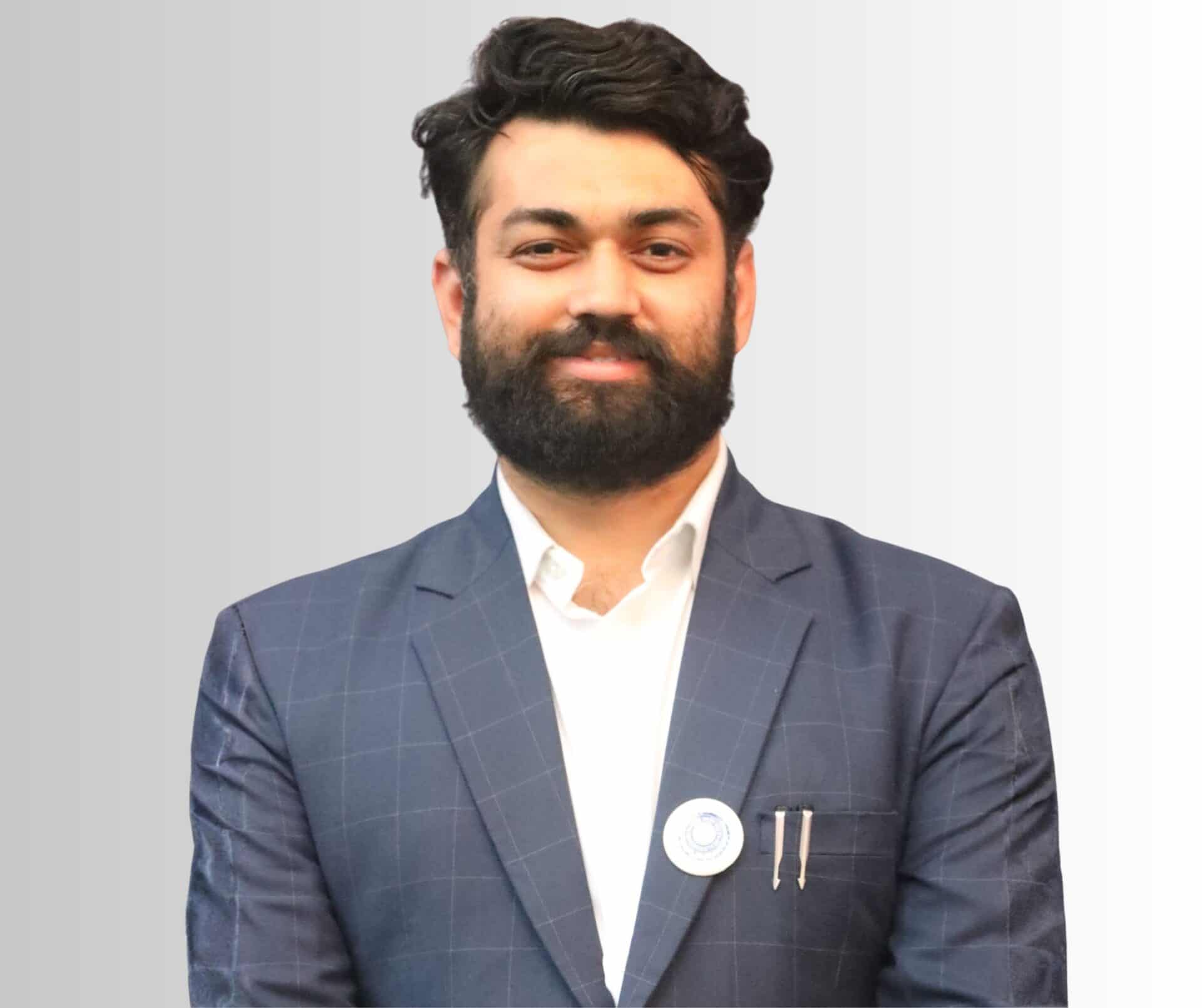 A bearded man wearing a white shirt and a blue jacket with a pin on the lapel.