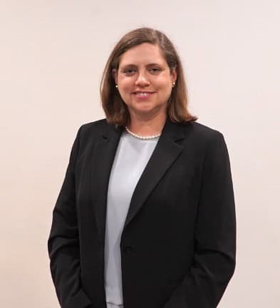 A person in a black blazer and white shirt stands against a plain background, smiling with their hands clasped.