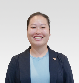 A person with tied-back hair, wearing a navy blazer over a light blue shirt, smiling at the camera against a plain light background.