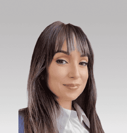 Portrait of a person with long dark hair and bangs, wearing a light-colored shirt and dark blazer, looking towards the camera against a plain background.