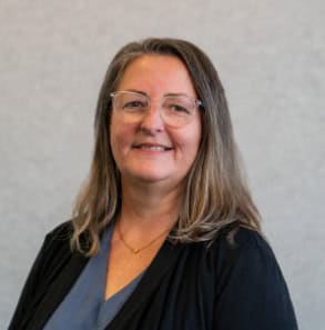 A woman with long brown hair, wearing glasses, a dark cardigan, and a gray top, stands against a plain light background, smiling at the camera.