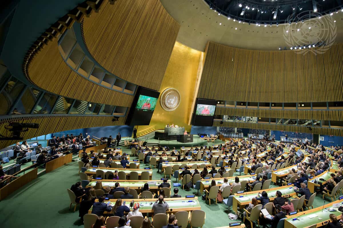 The United Nations General Assembly Hall in session with delegates seated at desks facing a central lectern.