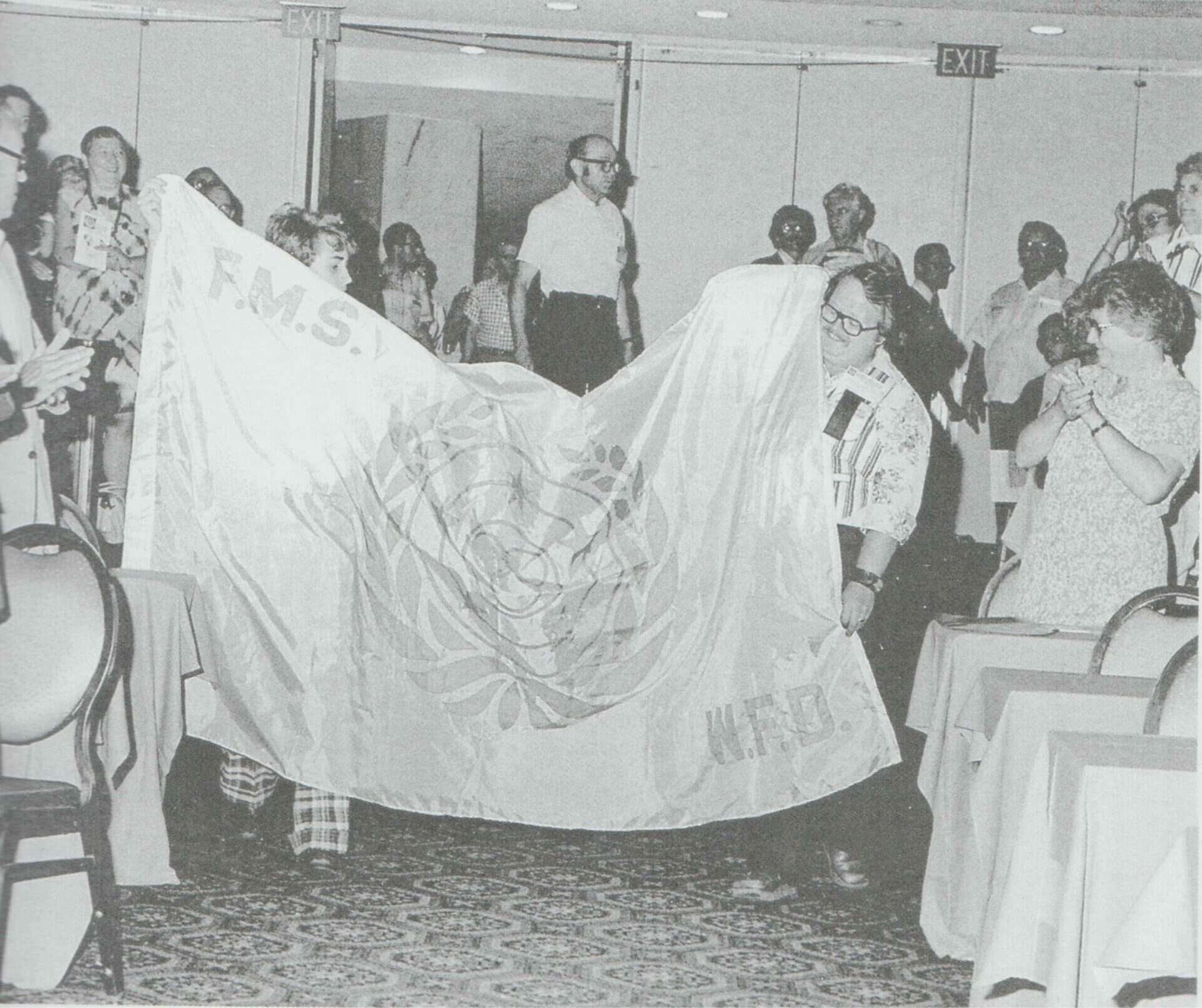 Two people hold up a large flag featuring the United Nations emblem, with the letters “F.M.S.” and “W.F.D.” visible, in a room with a group of onlookers.