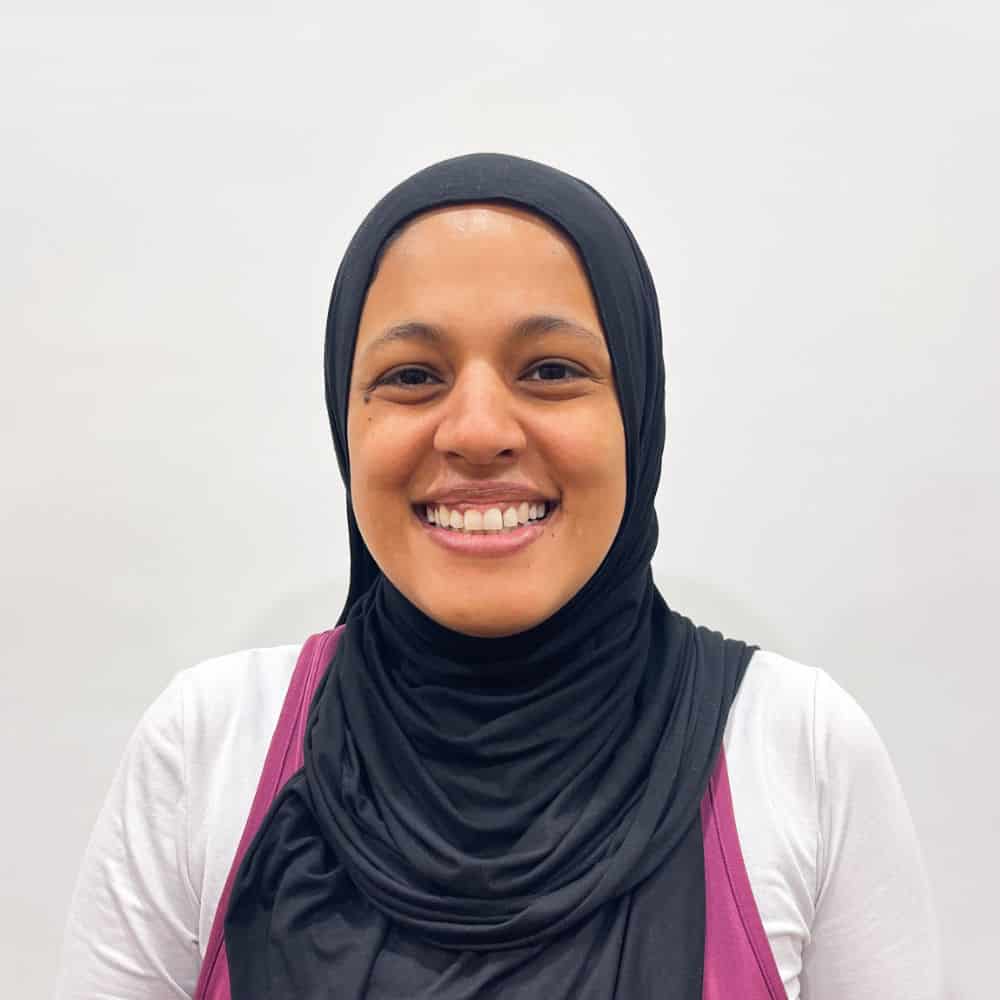 Person smiling, wearing a black hijab and white shirt, set against a plain background.