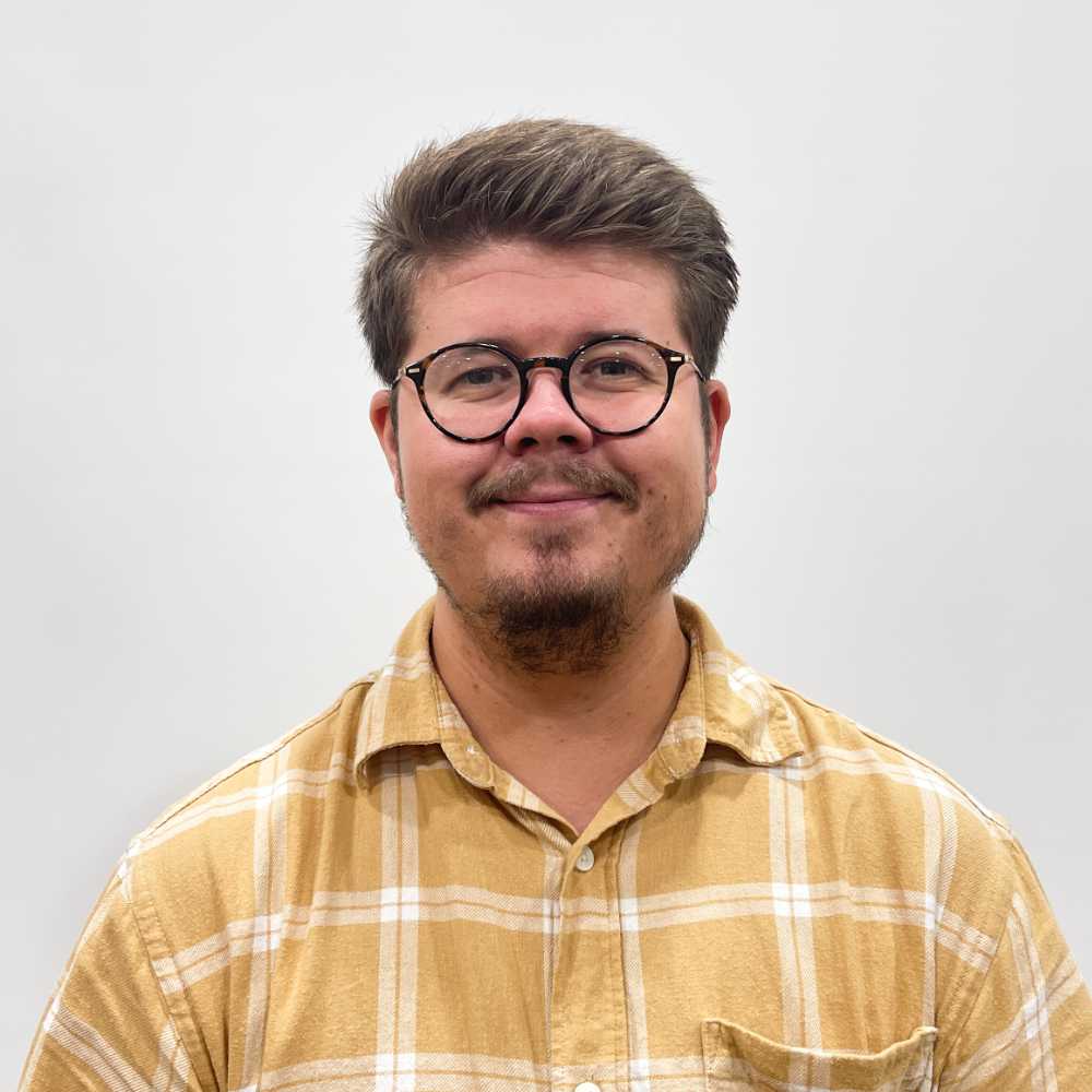 A person with short hair, glasses, and a beard, wearing a yellow plaid shirt, stands against a plain white background.