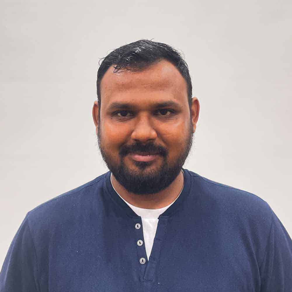 A person with short dark hair and a beard, wearing a dark blue shirt, stands against a plain background.