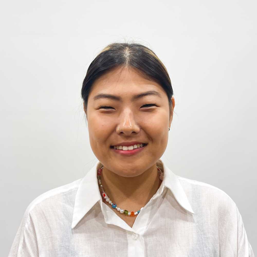 A person wearing a white shirt and a beaded necklace smiles against a plain white background.