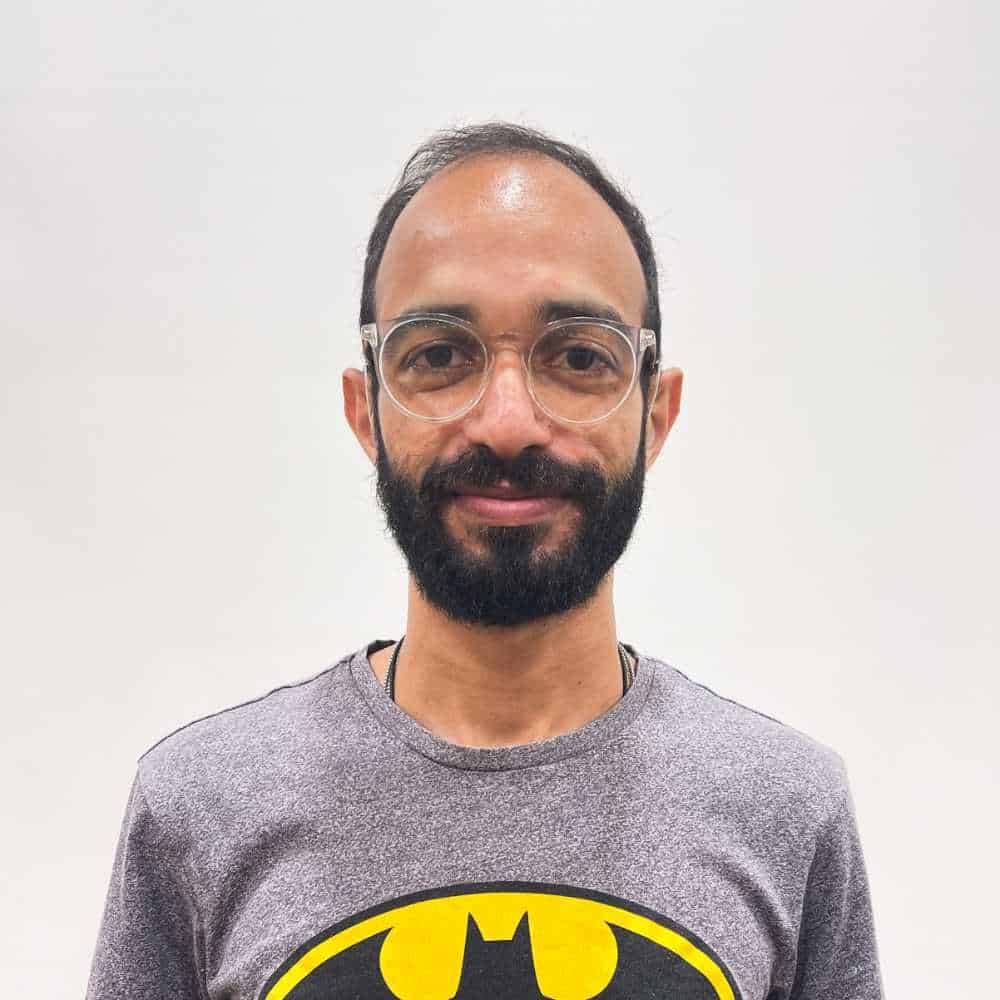 A person with glasses and a beard wears a gray Batman T-shirt, standing against a plain background.