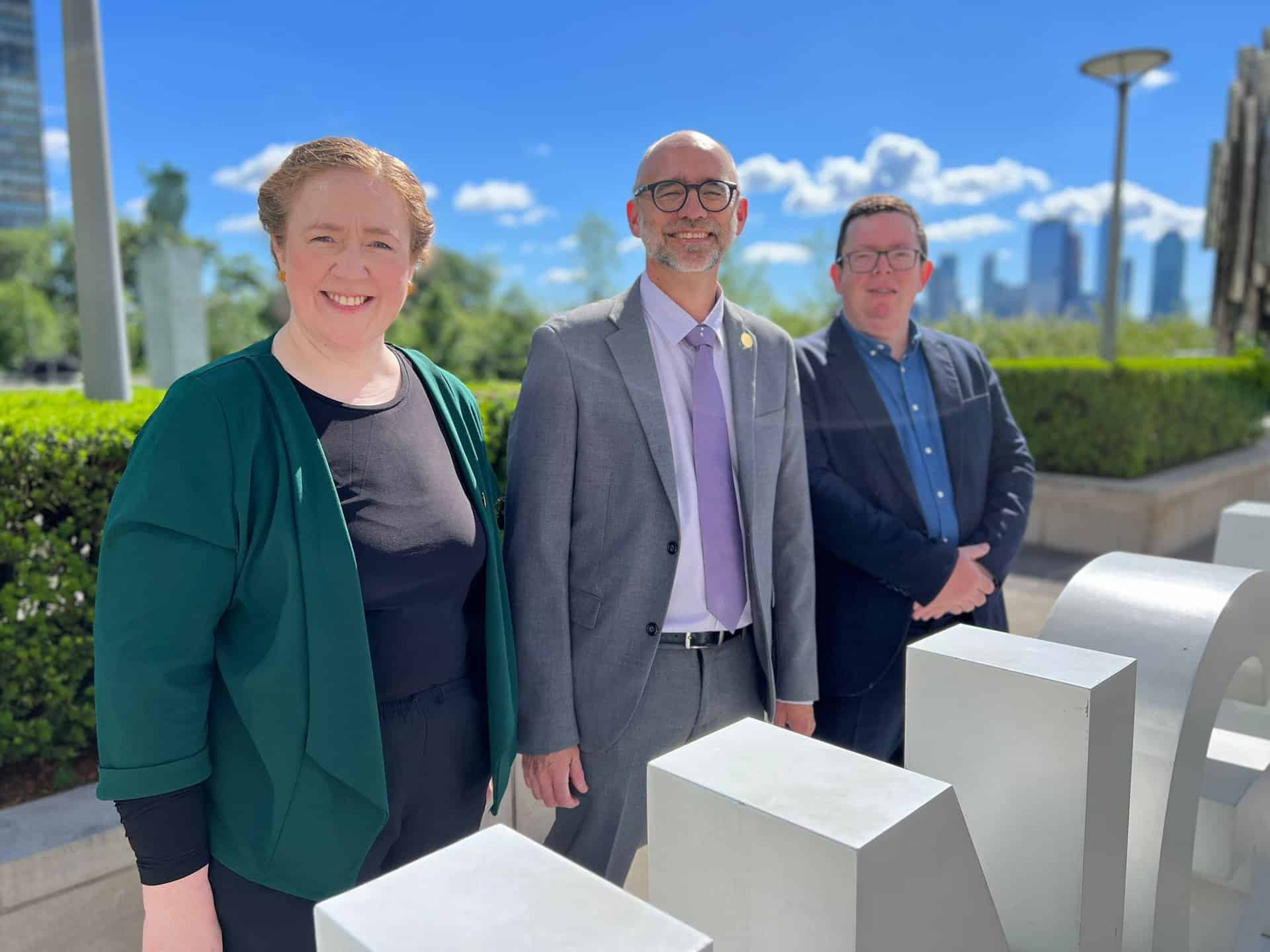 Three people standing outdoors on a sunny day, with a city skyline in the background. The woman on the left wears a green jacket, the man in the center wears a gray suit, and the man on the right wears a blue shirt.