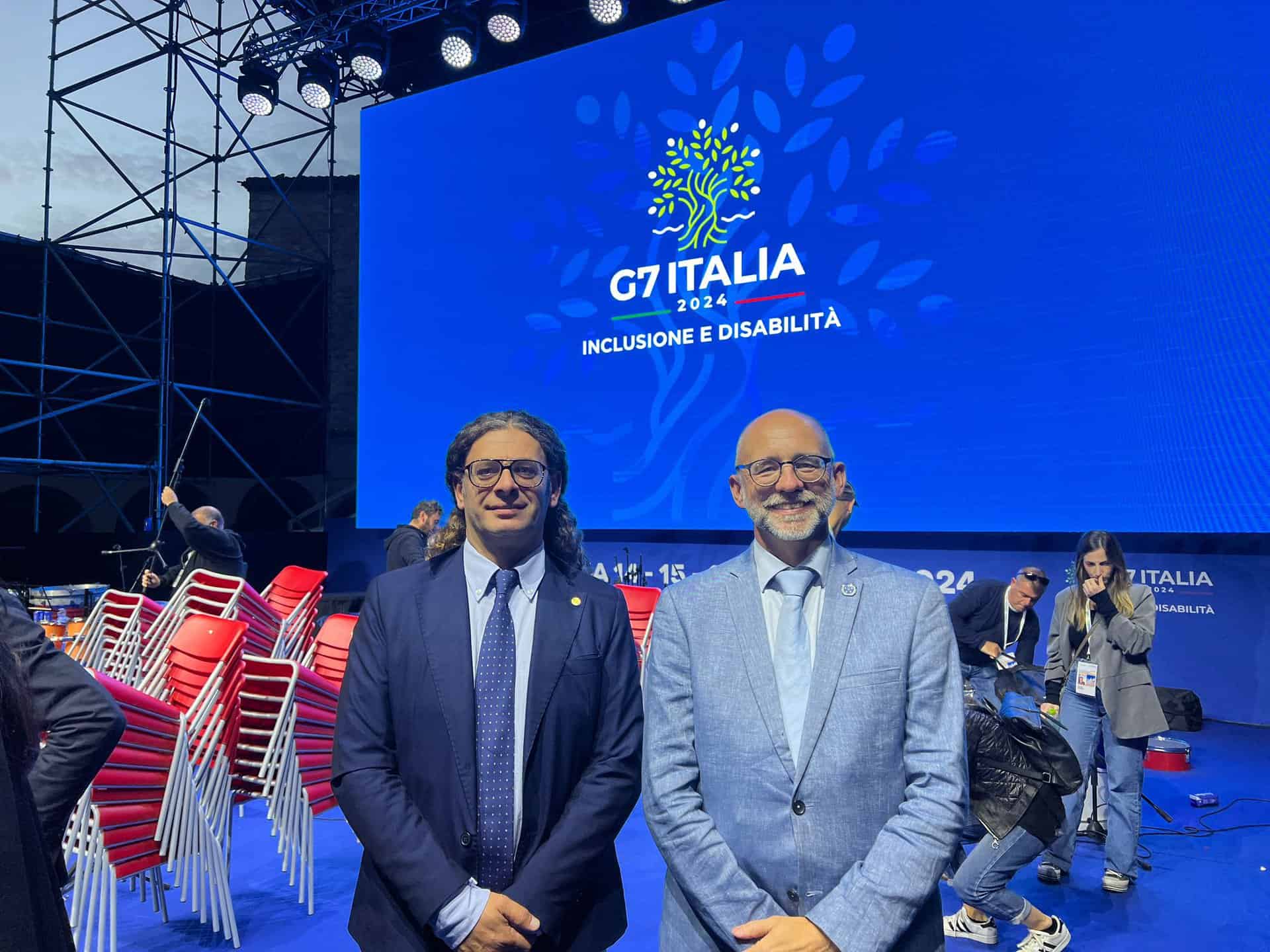 Two men in suits stand in front of a "G7 Italia 2024" backdrop displaying "Inclusione e Disabilità." Red chairs are stacked in the background.