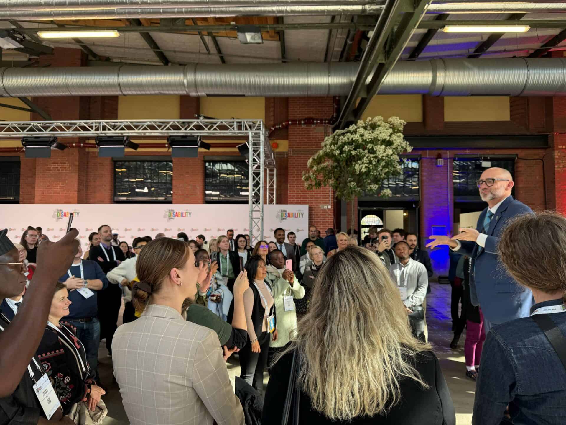 A speaker in a blue suit addresses a diverse group of seated and standing attendees in an industrial venue with banners and greenery.