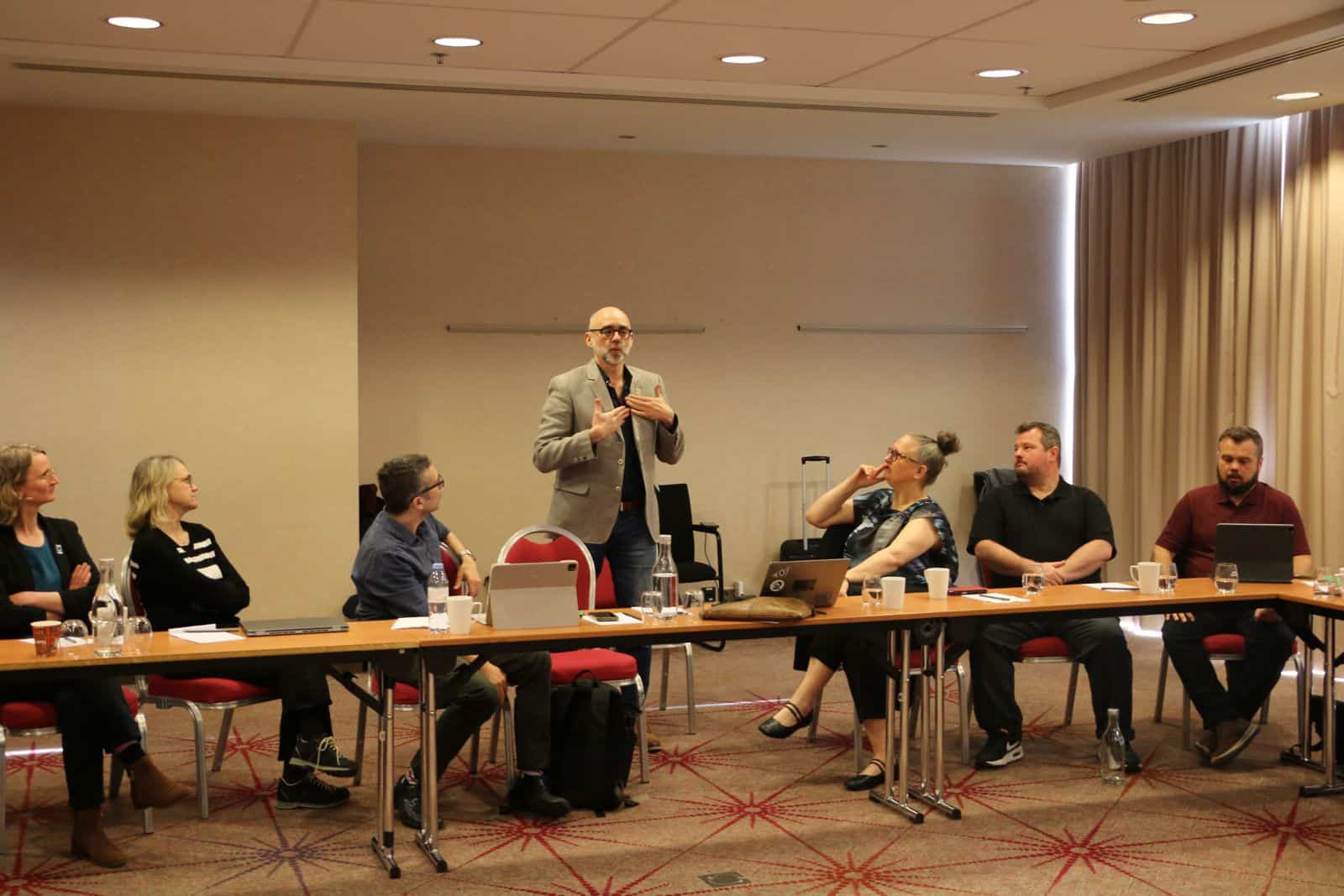 A man stands and speaks to a group of six seated people in a conference room, with laptops and papers on the table.