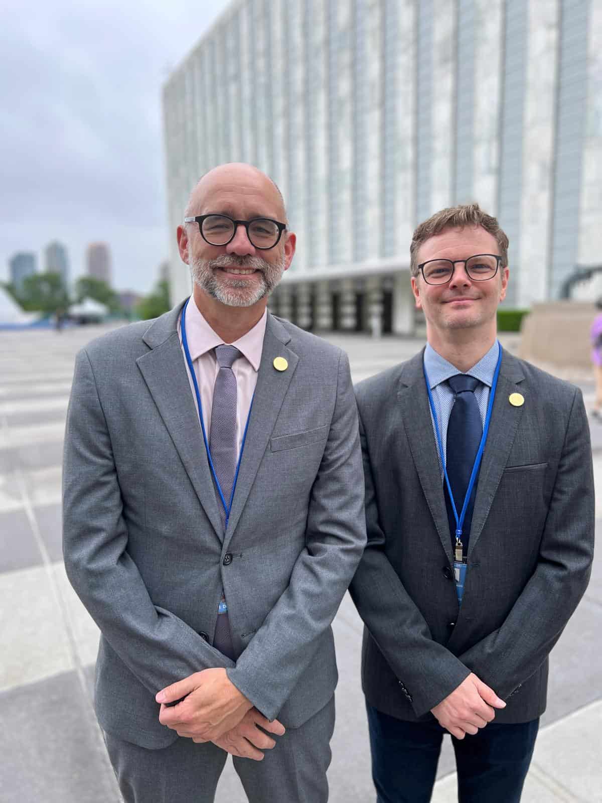 Two men in suits and lanyards stand outside a modern glass building, facing the camera and smiling.