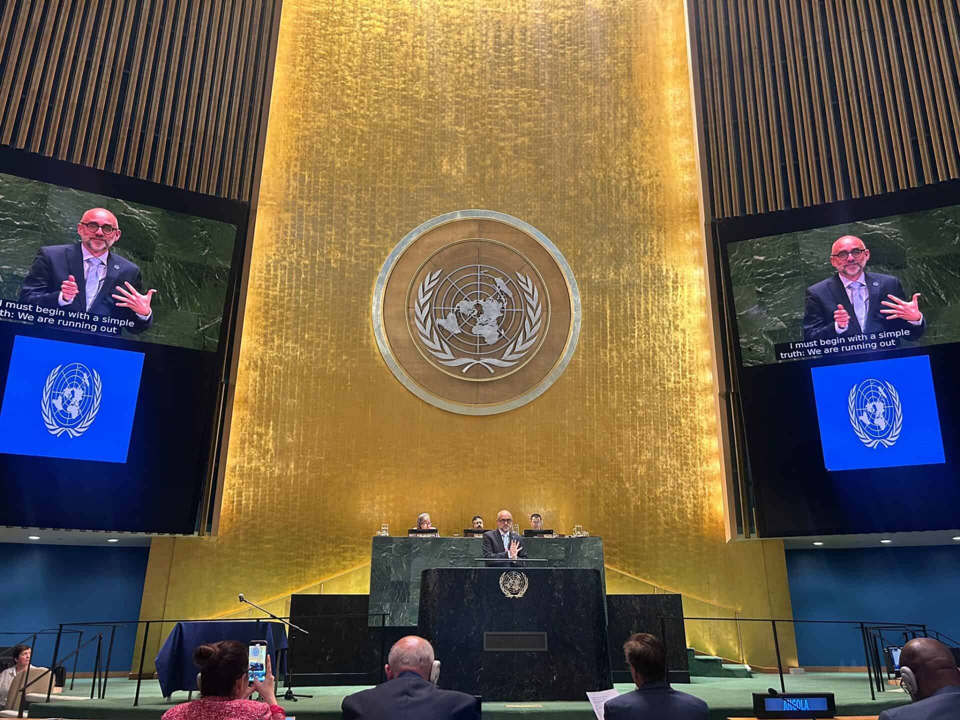 A speaker addresses the United Nations General Assembly hall from a podium, with the UN emblem displayed behind and on side screens; attendees are seated and listening.