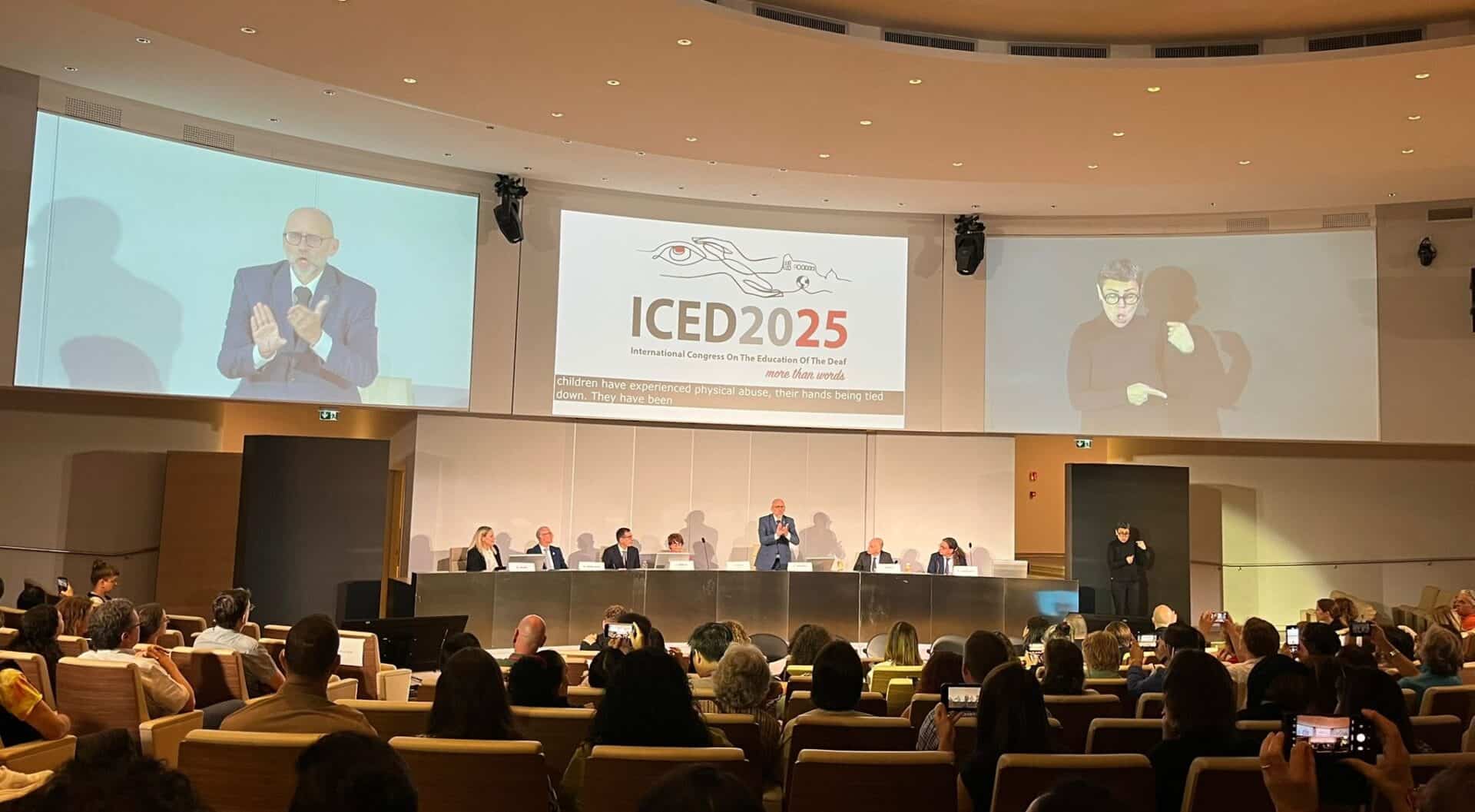 A conference panel sits on stage with a large screen displaying “ICED2025” and sign language interpreters present; audience members watch and record with phones.