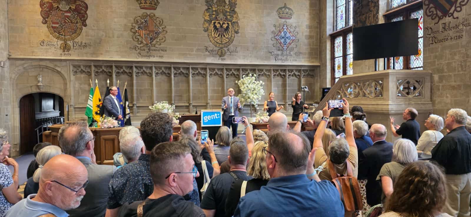 A large group of people attends a formal indoor event, with a speaker at a podium labeled "gent" in front of ornate stone walls and tall, stained glass windows.