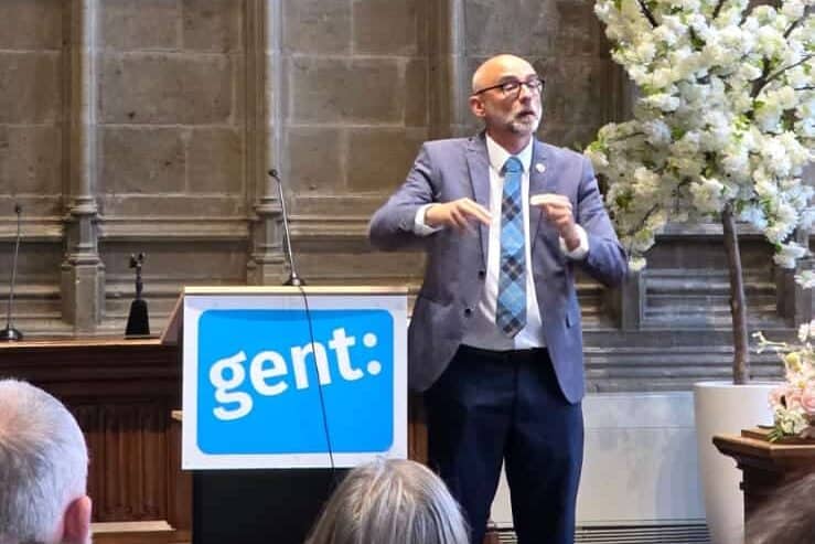 A man in a suit speaks at a podium labeled "gert:" in a historic hall with a large coat of arms and the date 1578 displayed on the stone wall behind him.