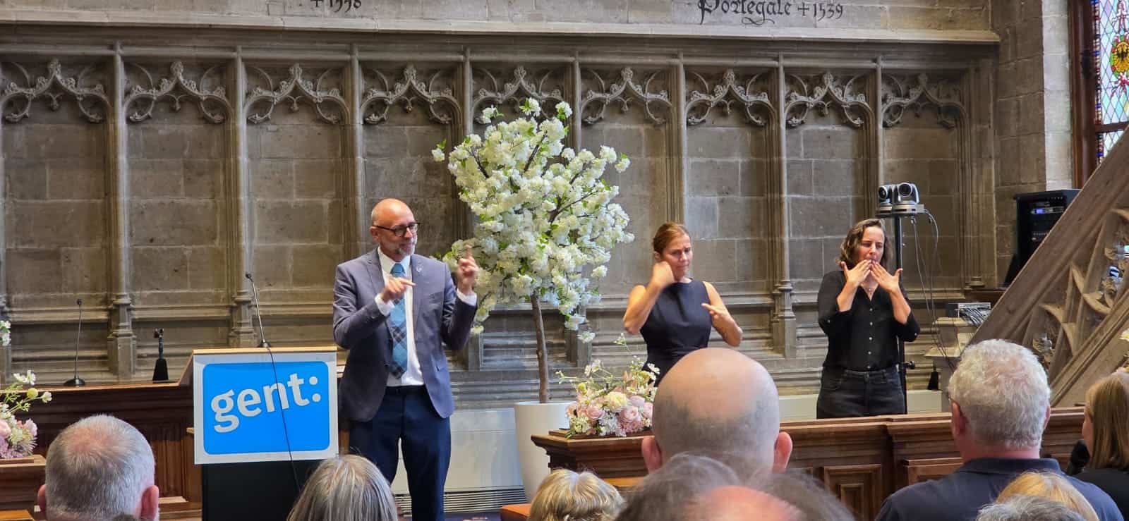 A man speaks at a podium labeled "gent:" while two sign language interpreters stand beside him in a church setting with an audience visible in the foreground.