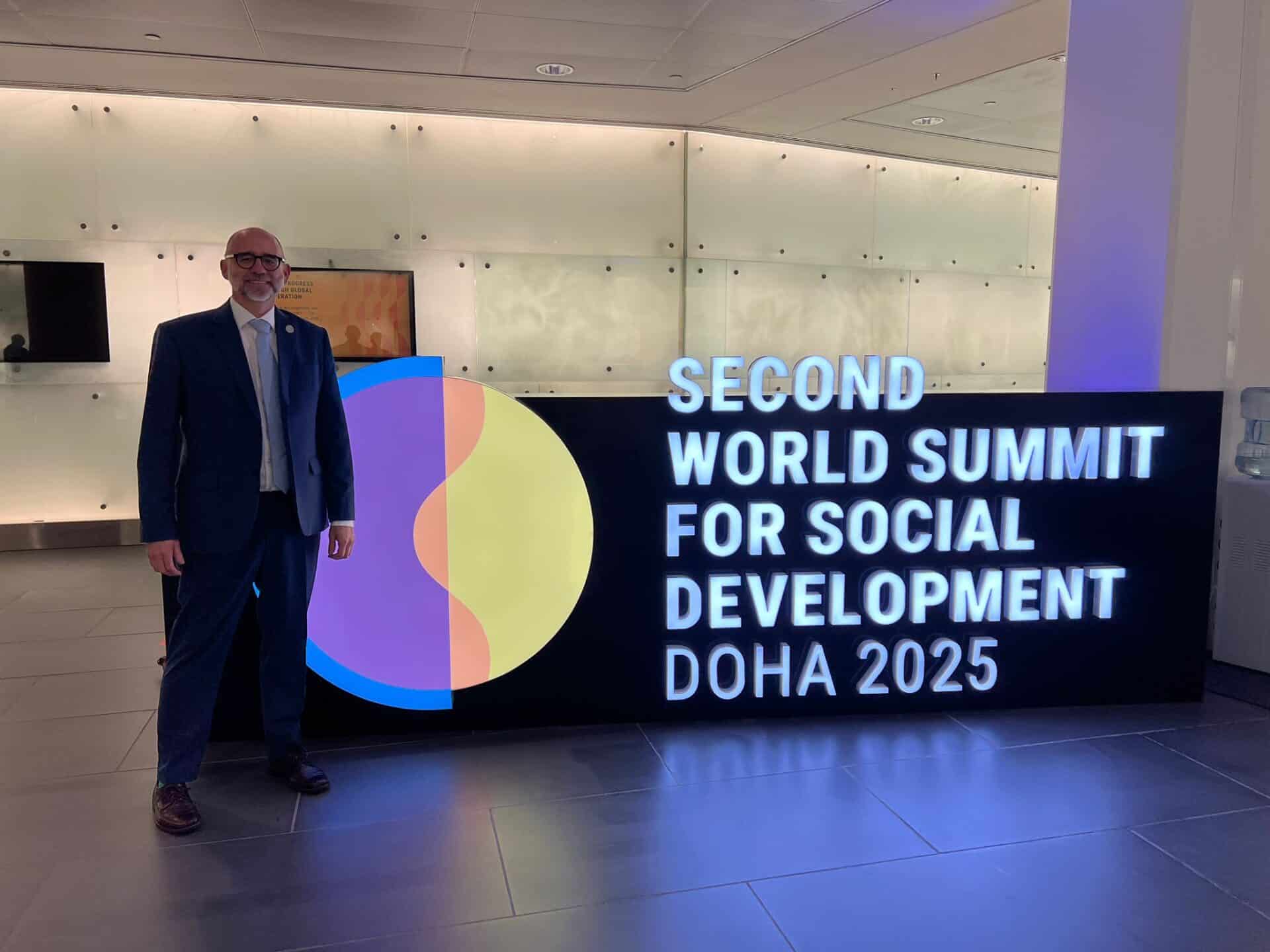 A man in a suit stands next to a sign reading "Second World Summit for Social Development Doha 2025" in a modern, well-lit indoor space.