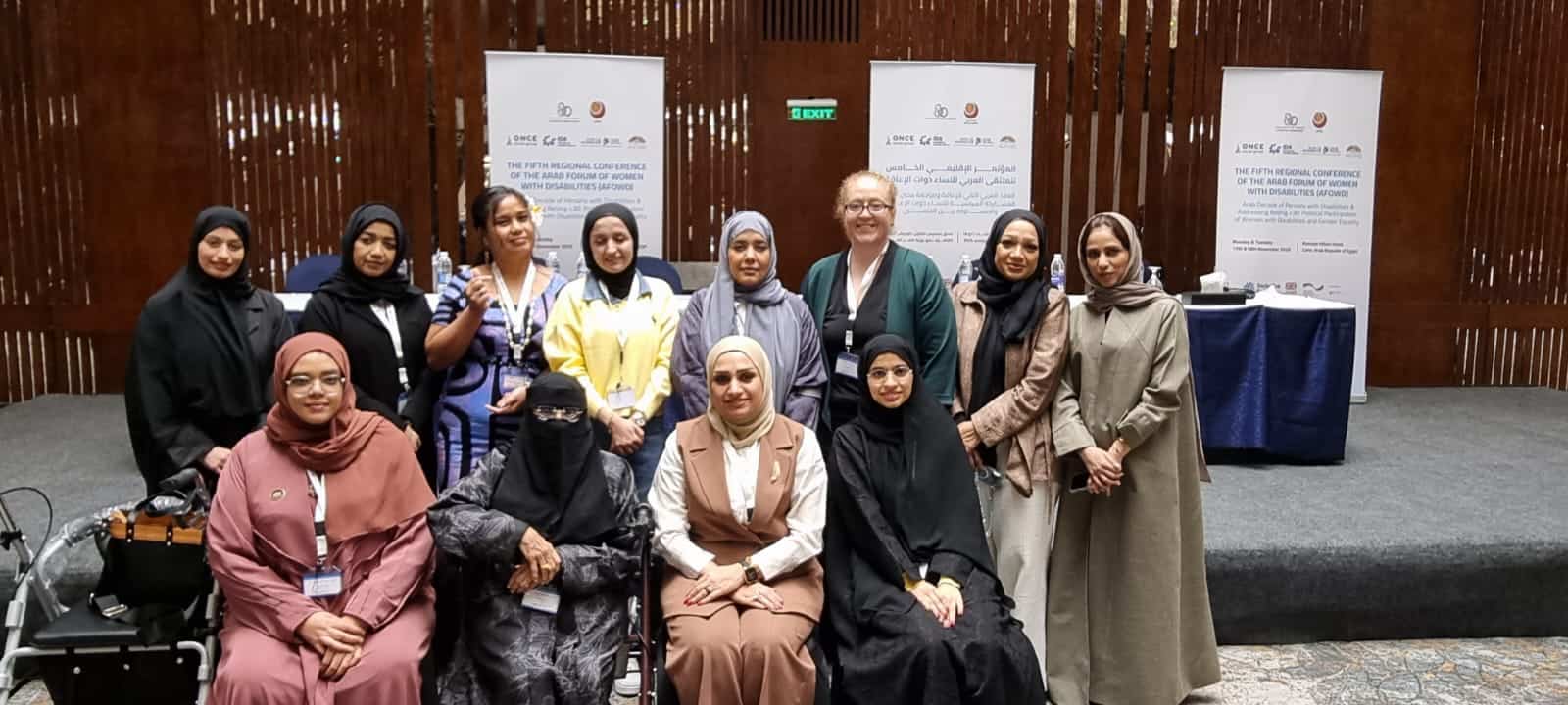 A group of women pose for a photo indoors in front of banners at a professional event or conference.