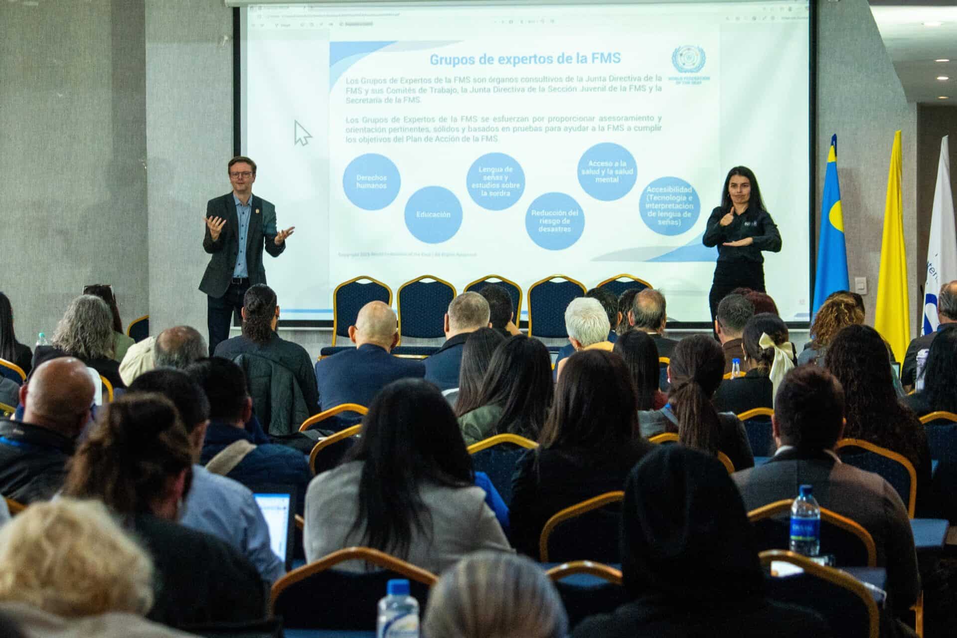 A man presents in front of an audience with a slide about expert groups projected on a screen; a woman stands beside him interpreting in sign language.