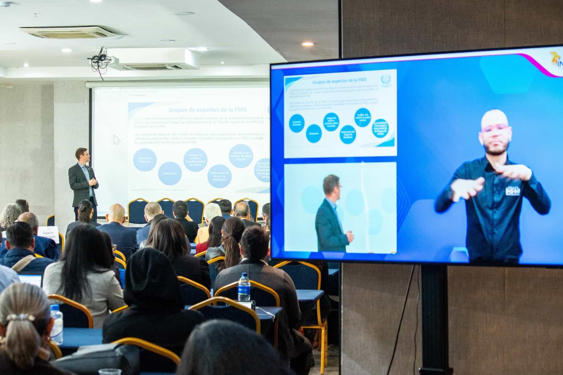 A man gives a presentation to an audience; a large screen displays a sign language interpreter and a duplicate of the presentation slides.