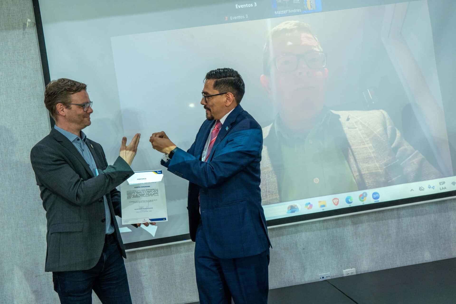 Two men in suits exchange a document and handshake gesture in front of a screen displaying a third man on a video call.
