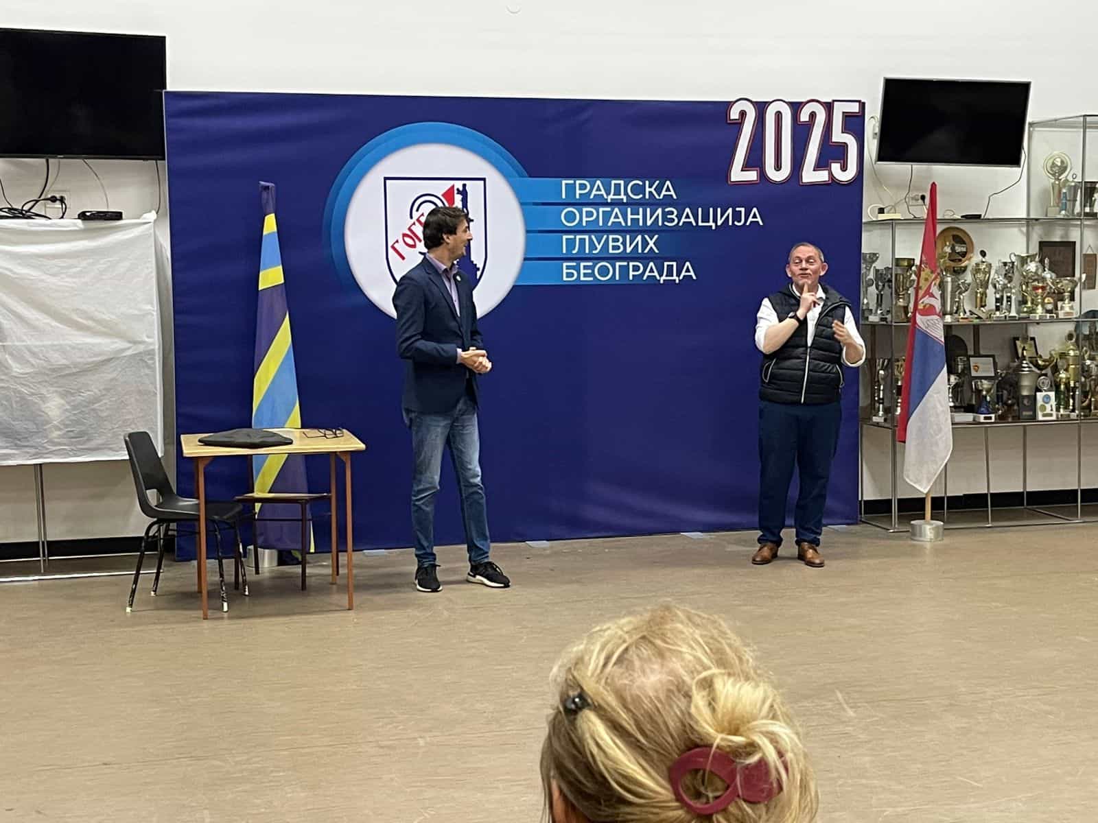 Two men stand on a stage in front of a blue backdrop with Serbian text, next to flags and trophies, addressing an audience.