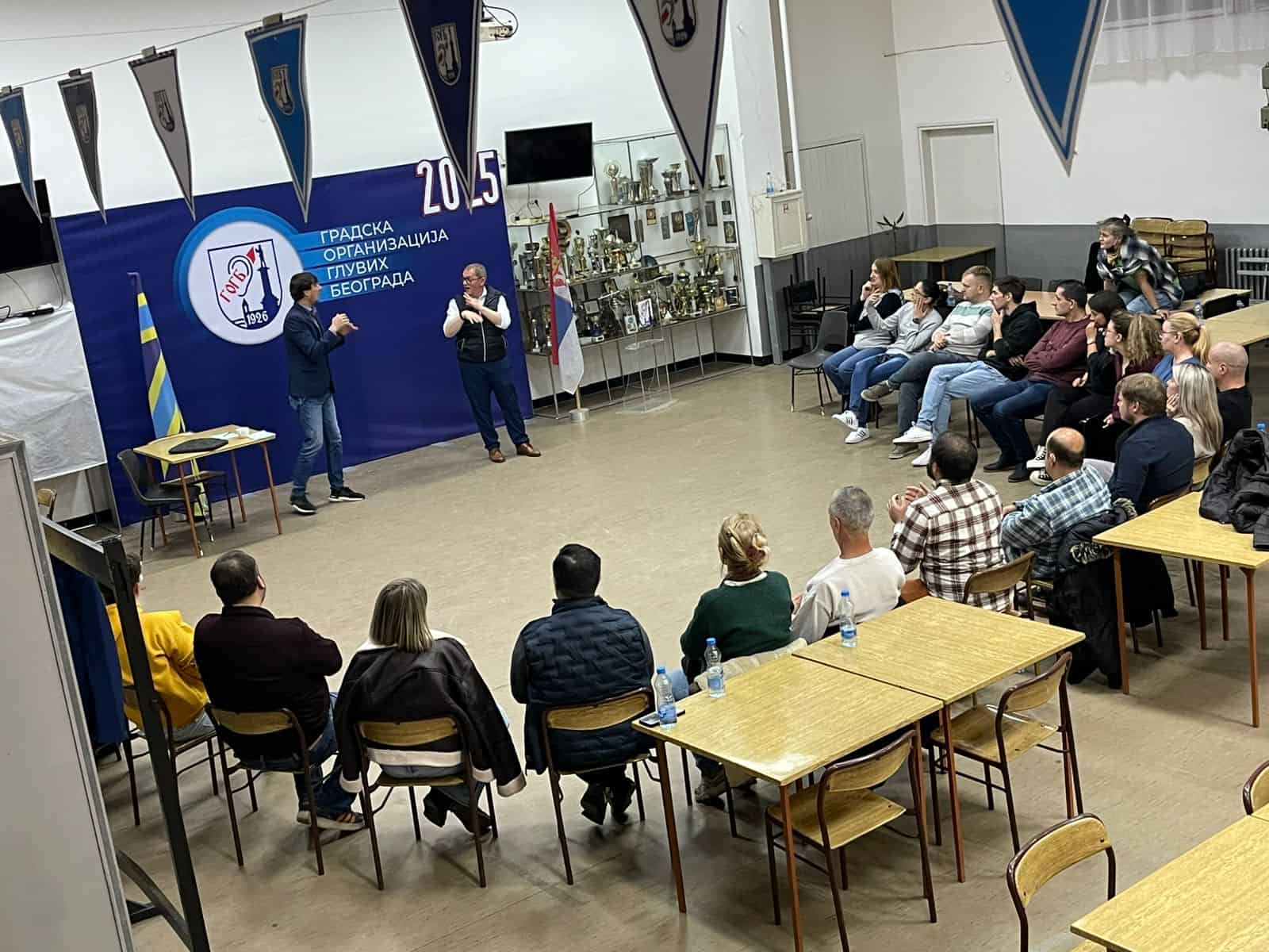 A group of people sit in a classroom, watching two presenters standing in front of a blue banner and a display of trophies.