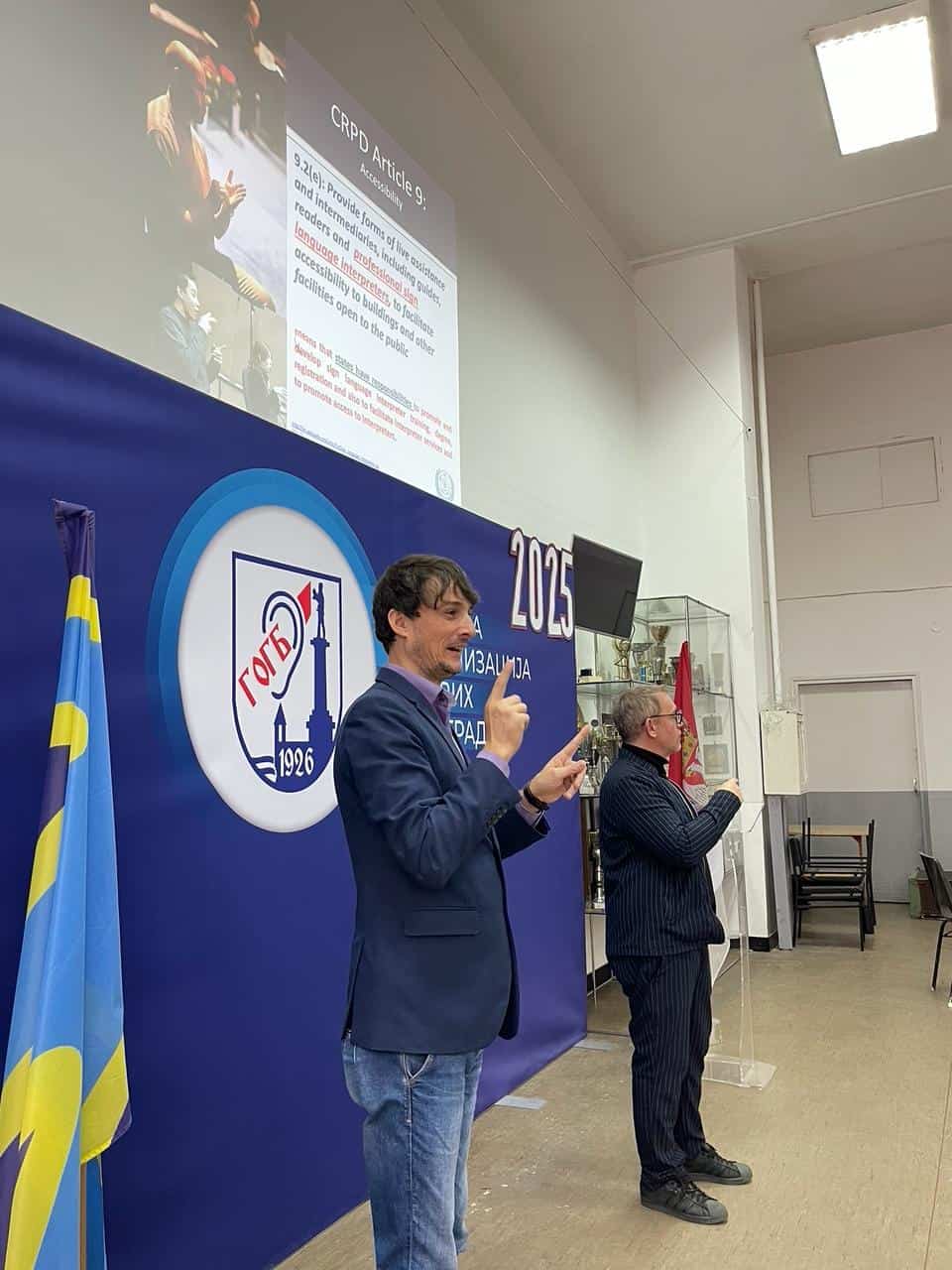 Two people stand in front of a blue backdrop with a logo, one interpreting in sign language. A presentation slide about CRPD Article 9 is projected on the wall behind them.
