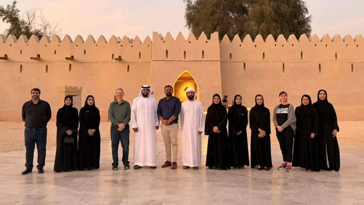 A group of people, men in traditional Middle Eastern attire and women mostly in black abayas, stand in front of a historic beige fort with crenellated walls at sunset.