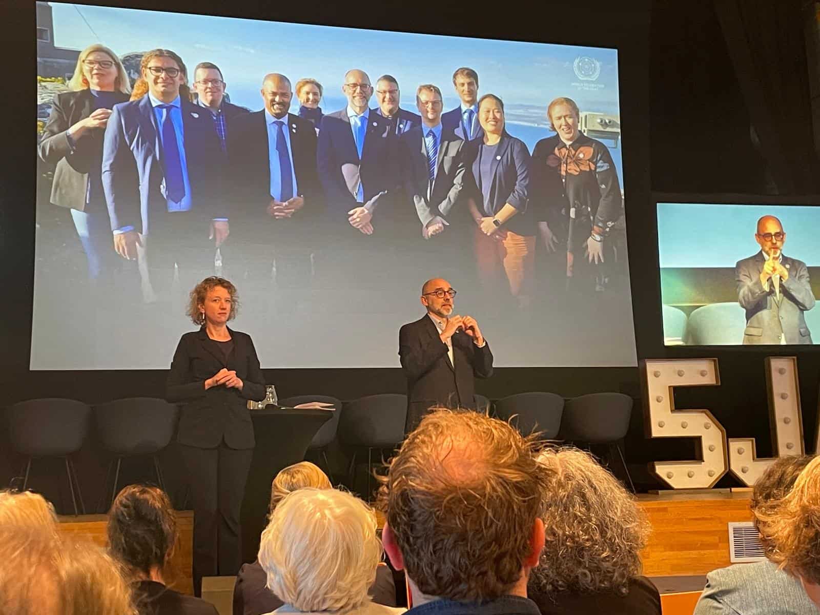 A man and a woman speak onstage at an event, with a projected photo of a group of people in suits displayed behind them. Audience members are seated in the foreground.
