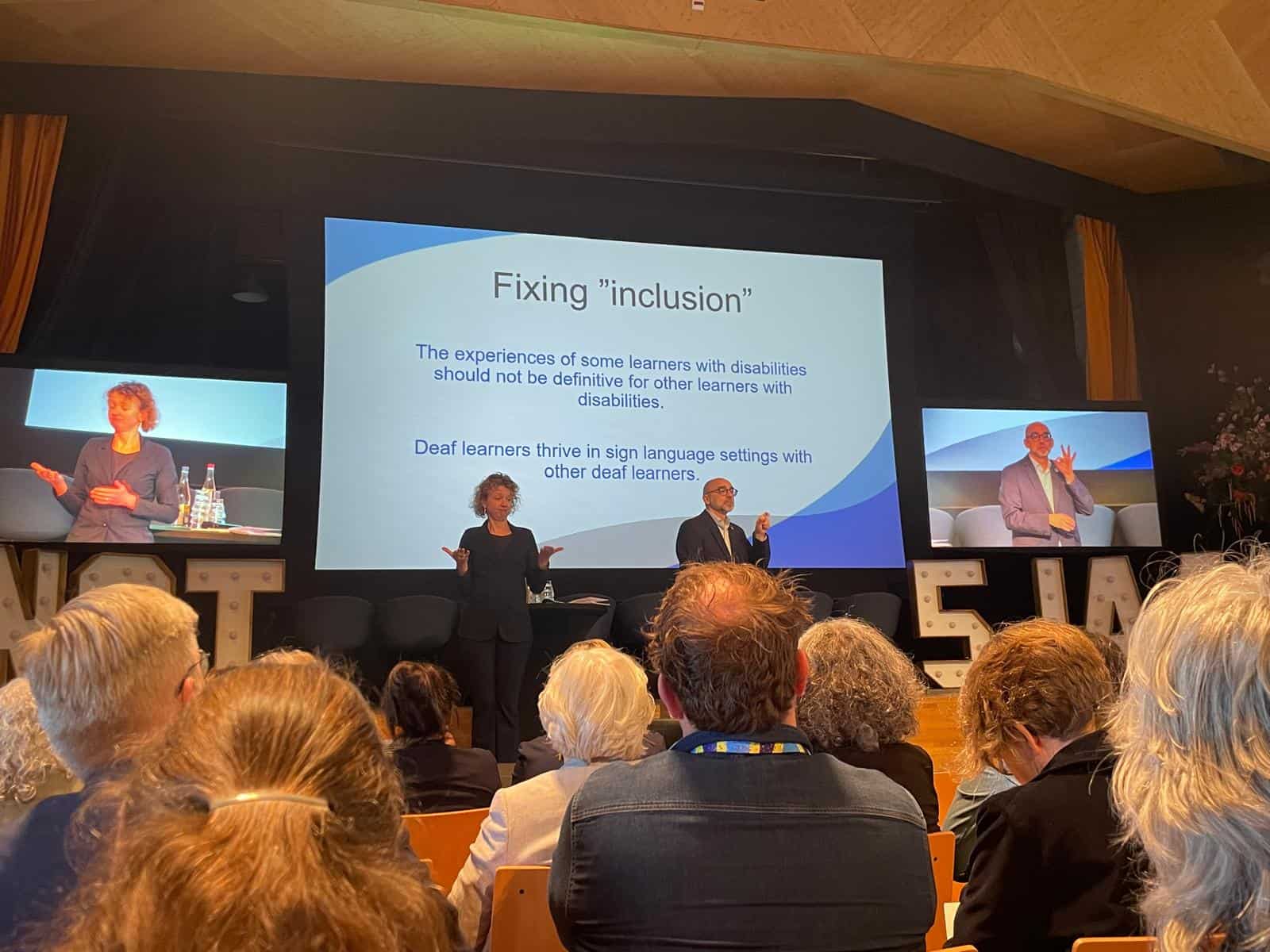 A presenter displays a slide on "Fixing 'inclusion'" to an audience in an auditorium, with two sign language interpreters on stage and the slide projected behind.